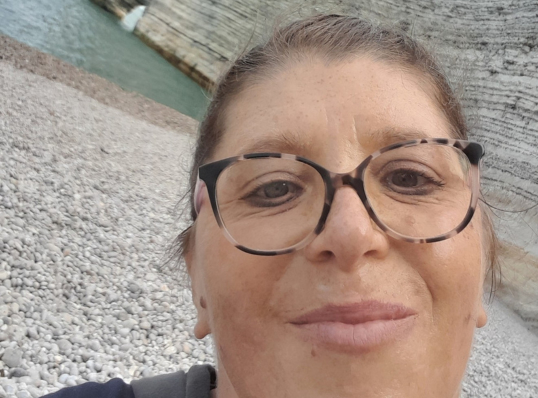 Portrait d’une femme portant des lunettes, souriante, en extérieur sur une plage de galets avec une falaise et de l’eau en arrière-plan.