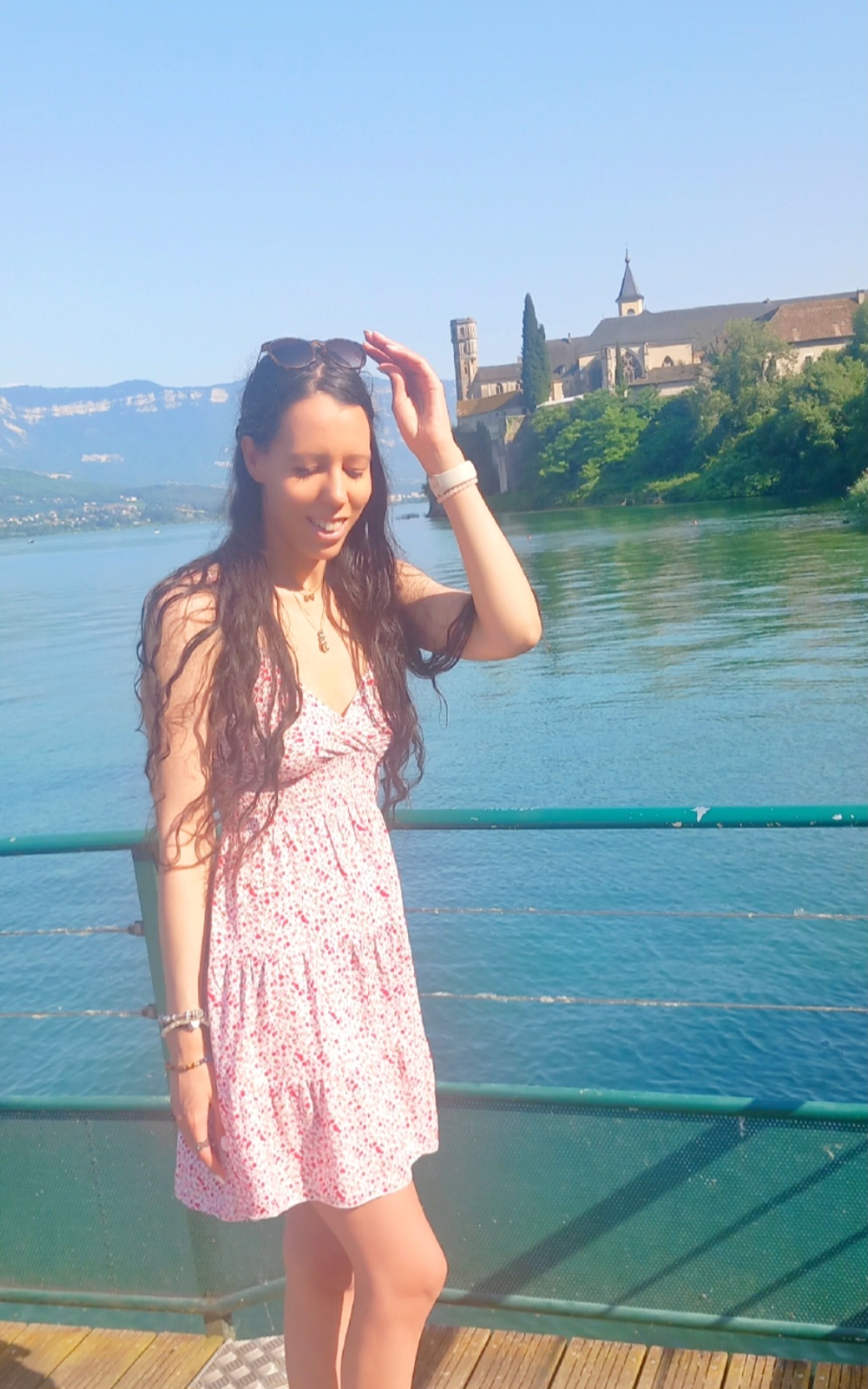 Eva souriante pose devant l’objectif sur un ponton. Derrière elle, une étendue d’eau reflète la lumière de l’été, et on aperçoit au loin un château. Elle porte une robe légère, évoquant une ambiance estivale.